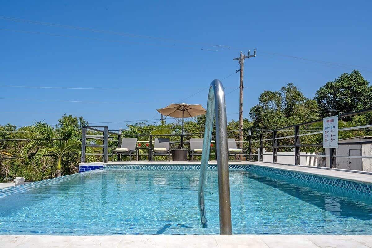 Una piscina azul cristalina con una escalera metálica, rodeada de sillas de descanso y una sombrilla, con árboles y un cielo despejado al fondo.
