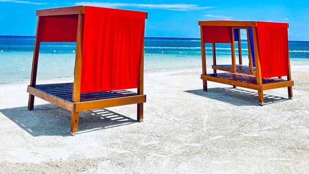 Deux chaises de plage en bois, surmontées de toiles rouges, se trouvent sur une plage de sable fin, avec une eau et un ciel d’un bleu limpide.