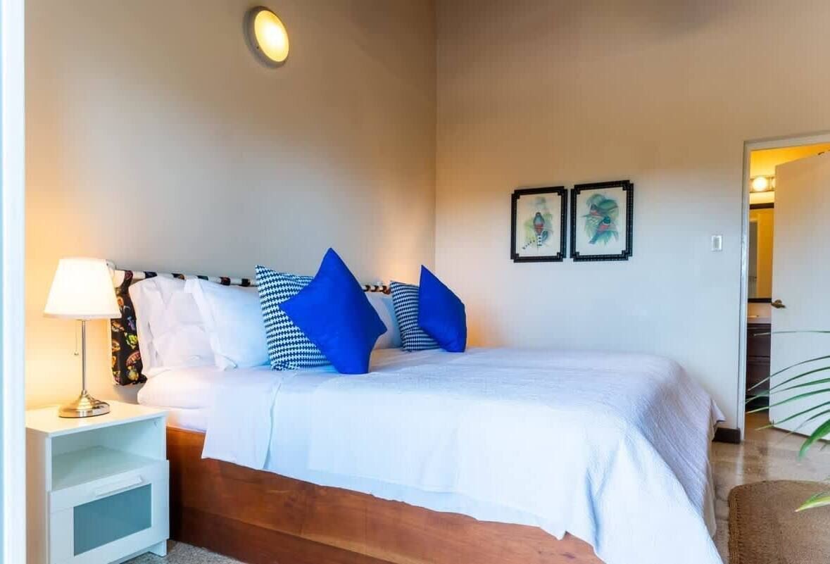 Un lit bien fait, avec du linge de lit blanc et des oreillers bleus, une table de chevet avec une lampe, et deux tableaux encadrés accrochés au mur.