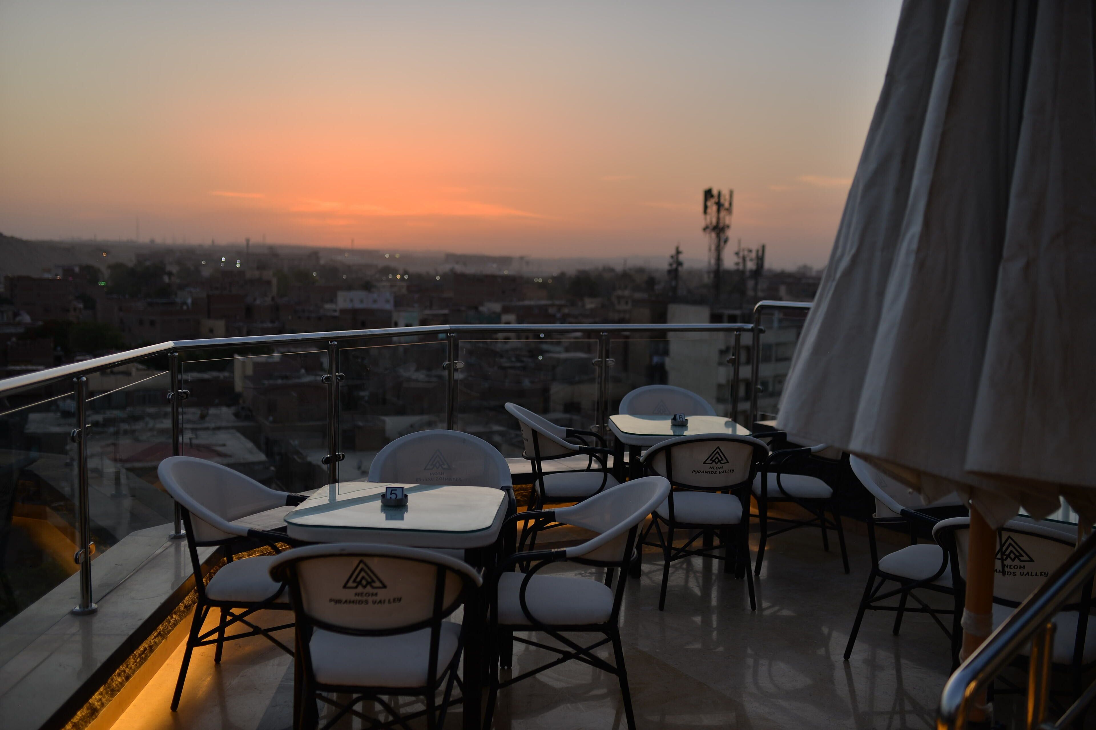A terrace with tables and chairs, a view of the cityscape, and a sunset.