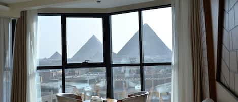A hotel room with large windows offering a view of pyramids. There are two chairs and a small table in the room.