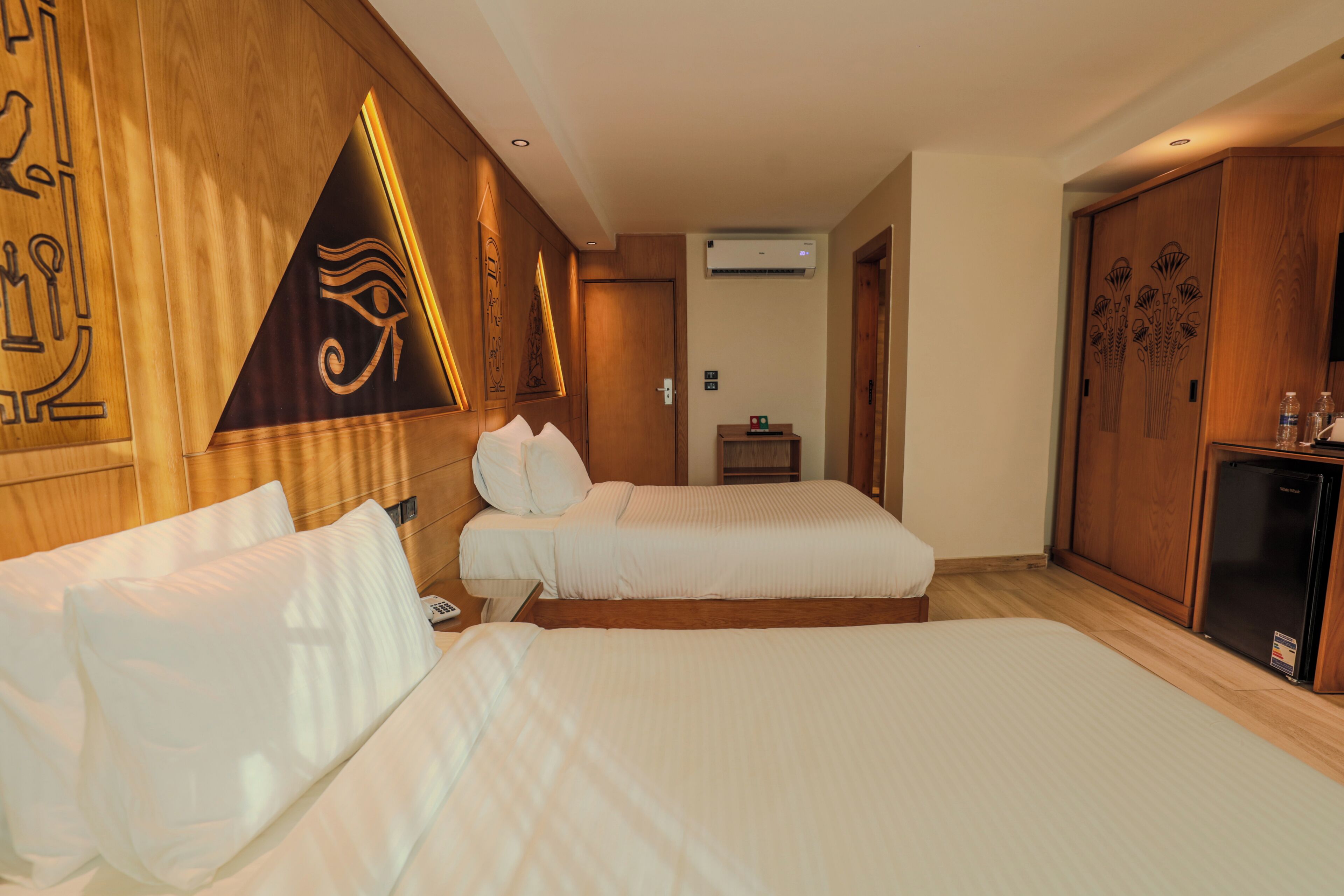 A hotel room with two beds, wooden headboards, a minibar, and an air conditioning unit.