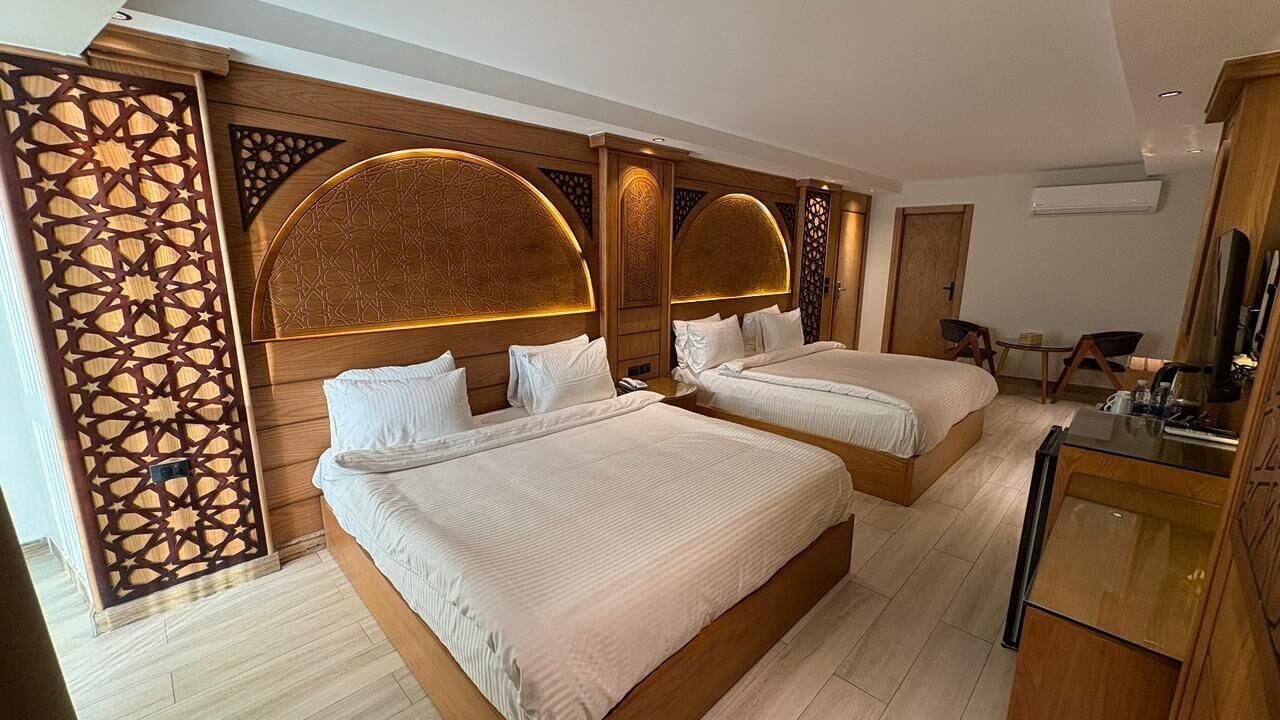 Habitación de hotel con dos camas, cabecero de madera con diseños elaborados, un escritorio pequeño y un televisor.