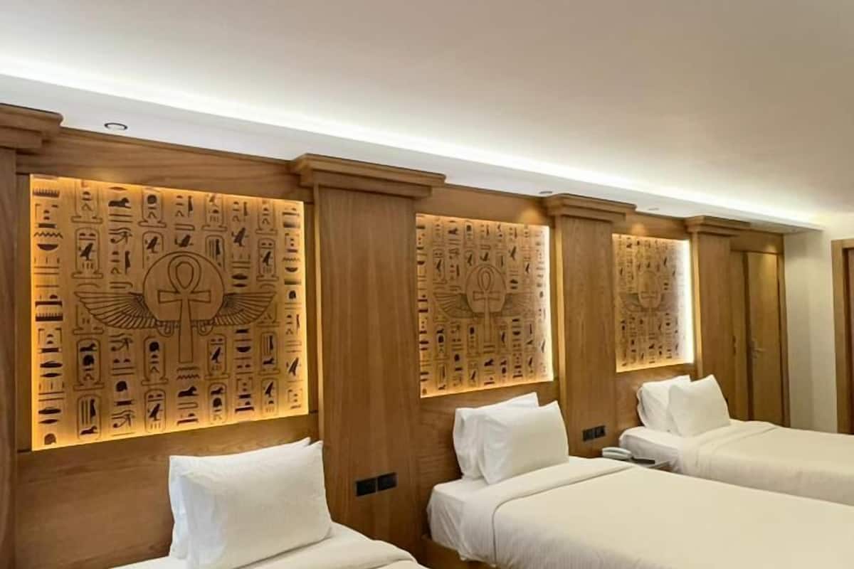 A hotel room with three beds, wooden headboards, and wall panels with hieroglyphs.