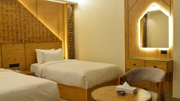 Habitación de hotel con dos camas, cabecera de madera con jeroglíficos, mesita de noche con superficie de cristal, mesa redonda de madera con una silla y un espejo con lámpara geométrica.
