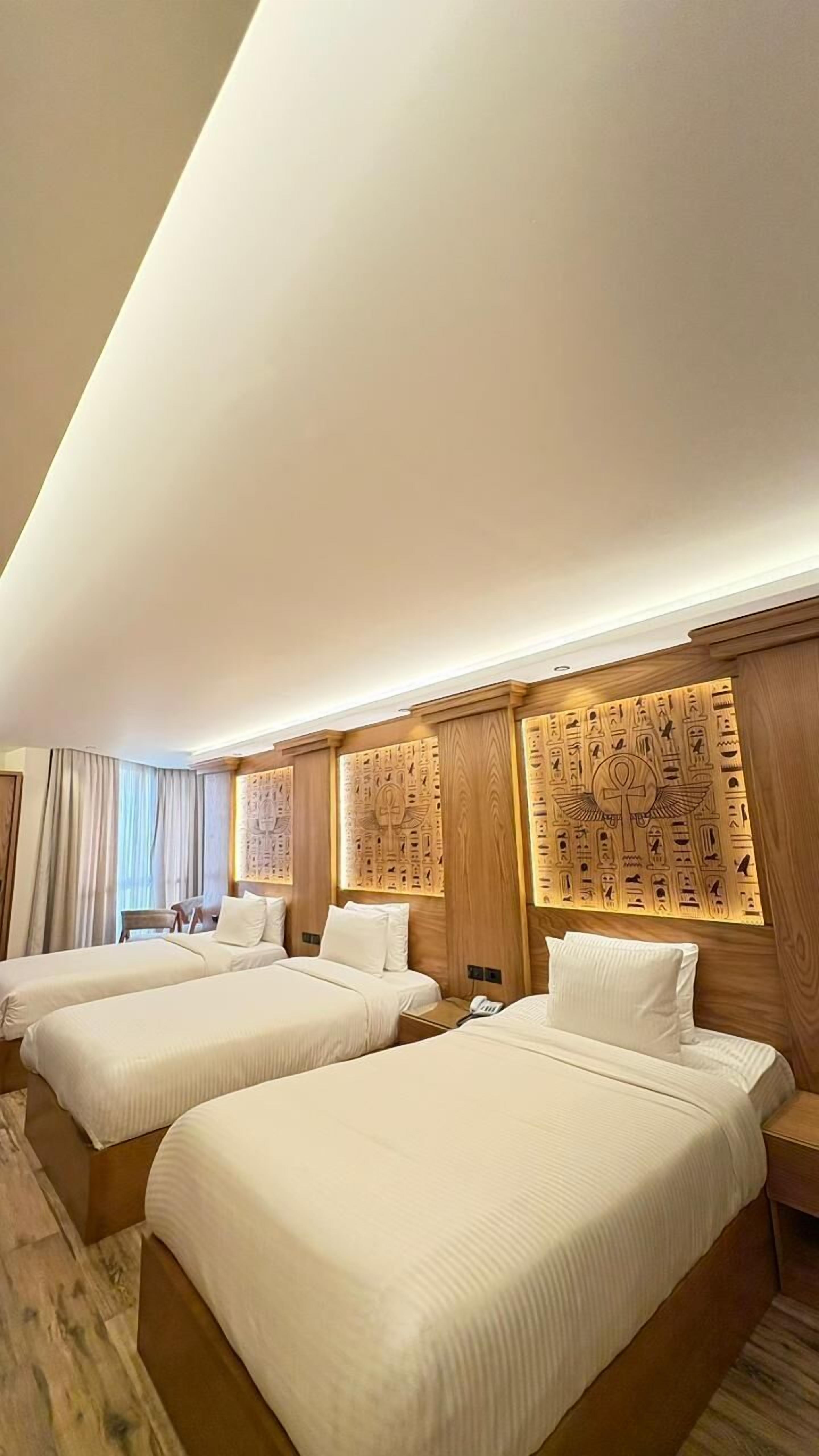 Ein Hotelzimmer mit zwei Betten, einem Holzkopfteil mit Hieroglyphen, einem Schreibtisch und einem Stuhl.