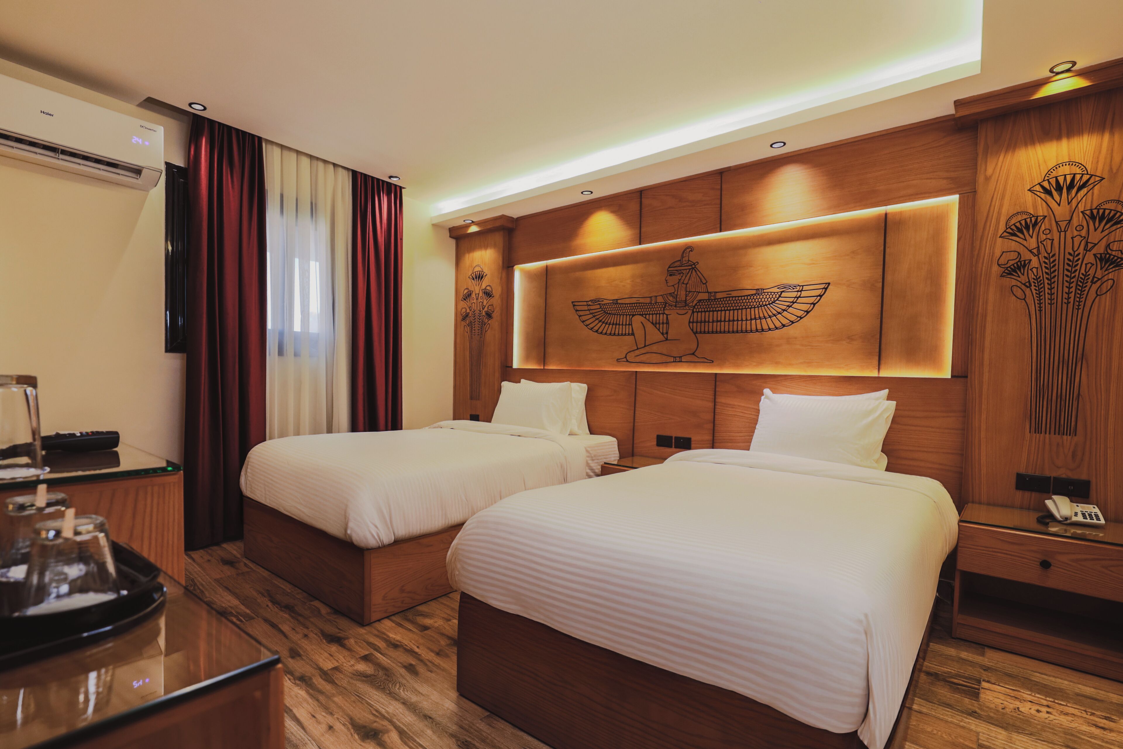 A hotel room with two beds, a wooden headboard with an Egyptian-themed design, a minibar, and a desk with a lamp.