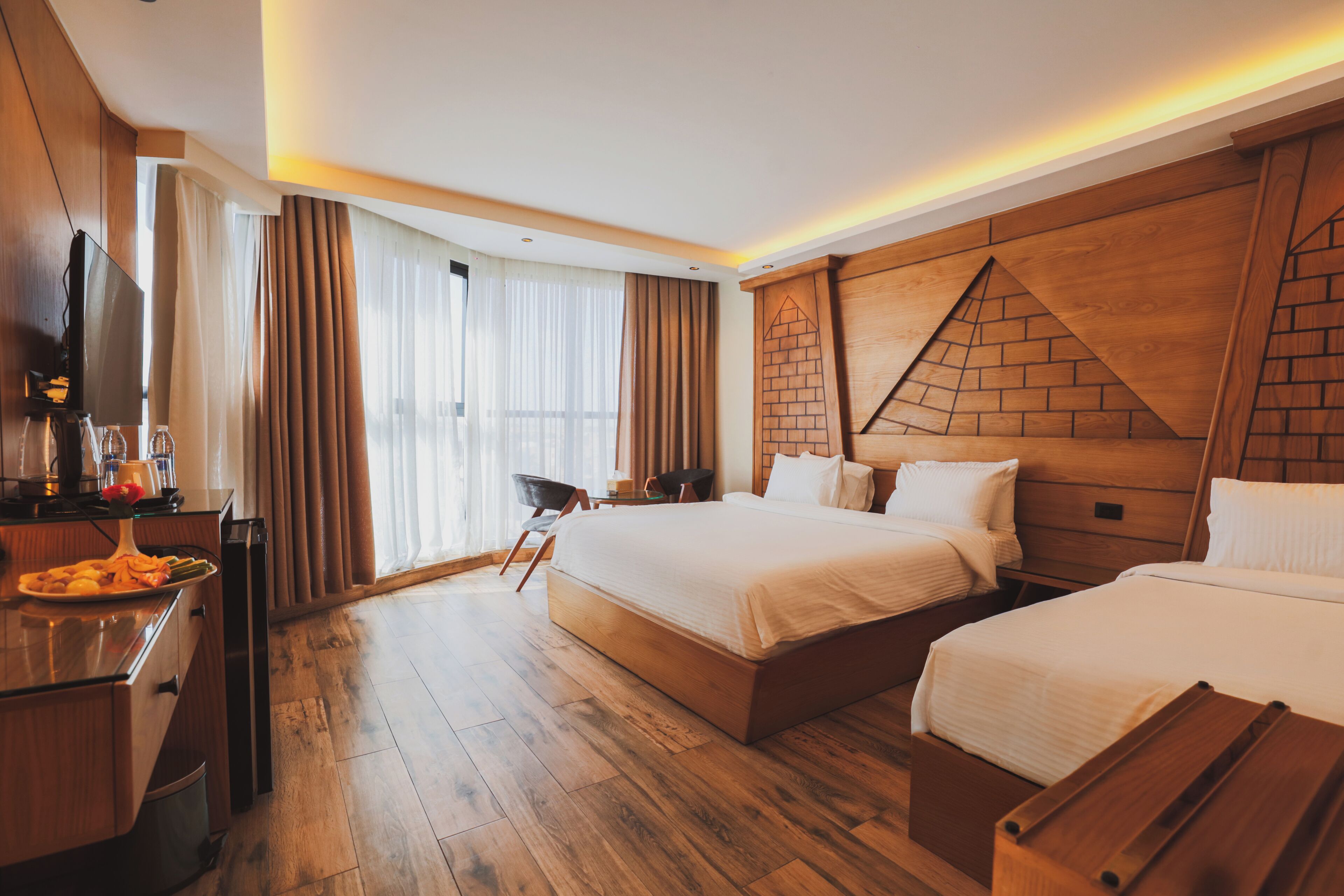 A hotel room with two beds, a wooden headboard, a flat-screen TV, a desk with a coffee maker, and a dining table with a plate of food.