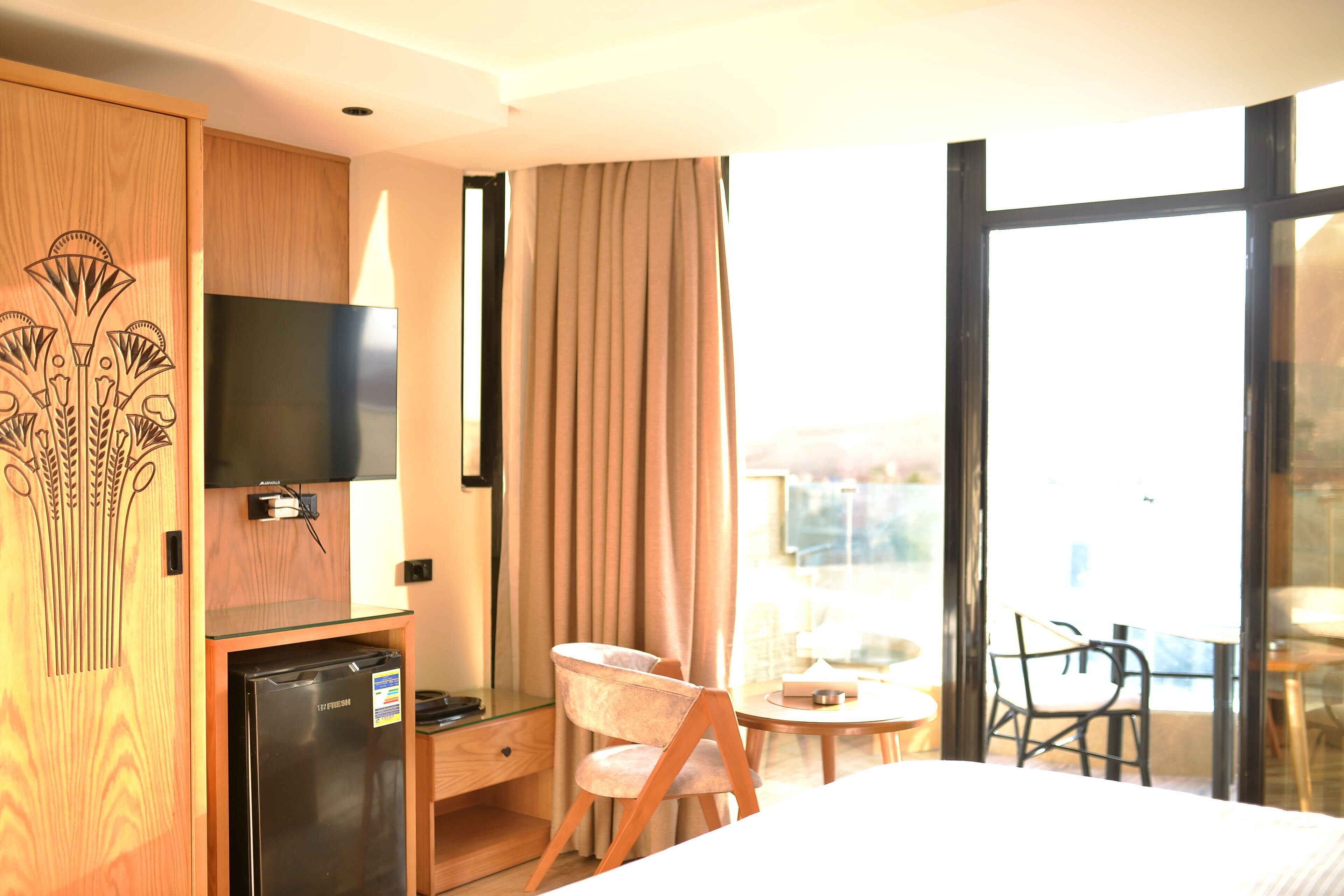 A hotel room with a bed, a desk with a television, a refrigerator, a chair, and a balcony with a table and chairs.