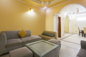 A living room with a sofa, ottoman, and glass coffee table. There is a dining area with a table and chairs in the background.