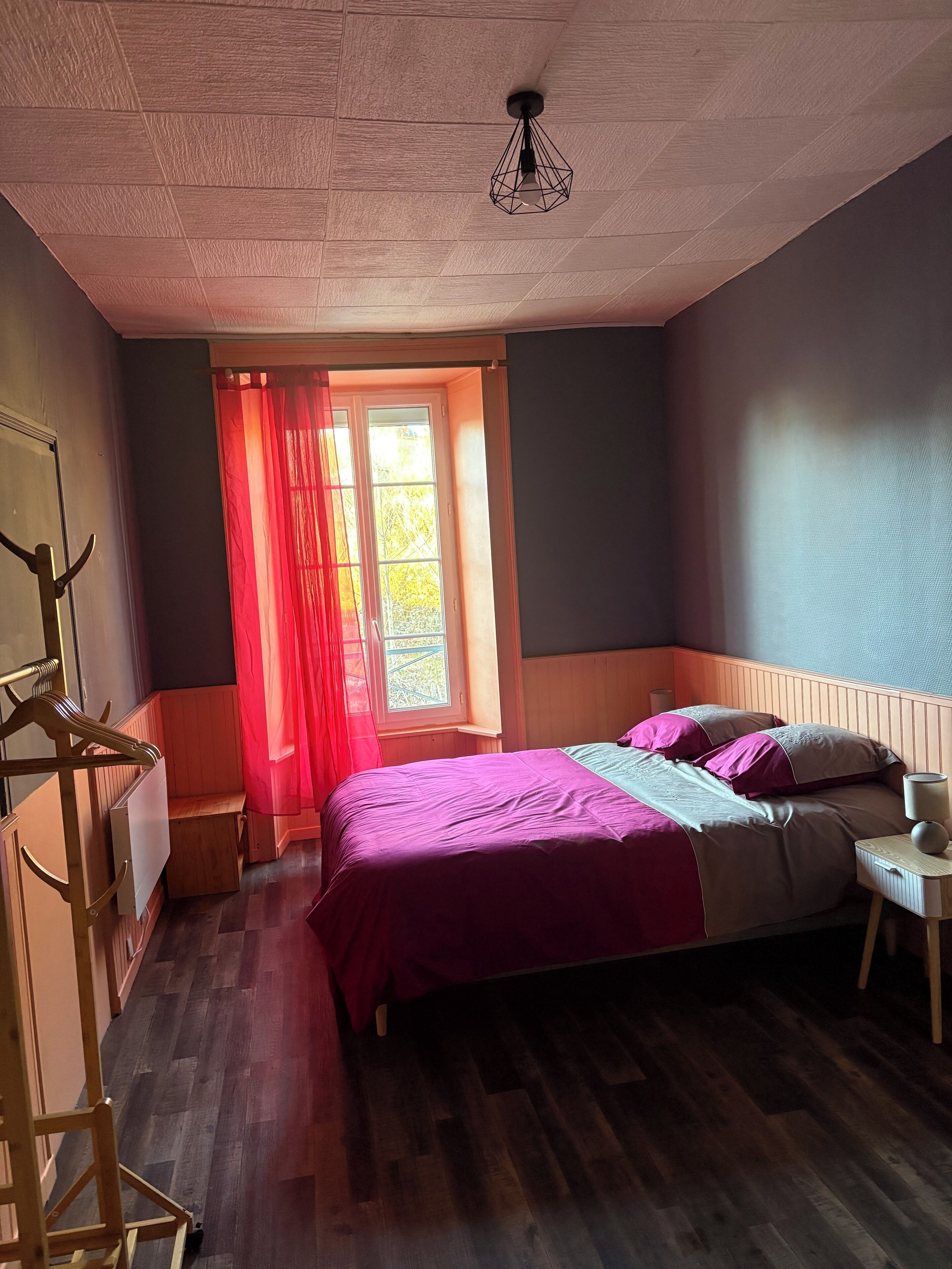 Un dormitorio con cama, ventana con cortinas, radiador y lámpara colgante.