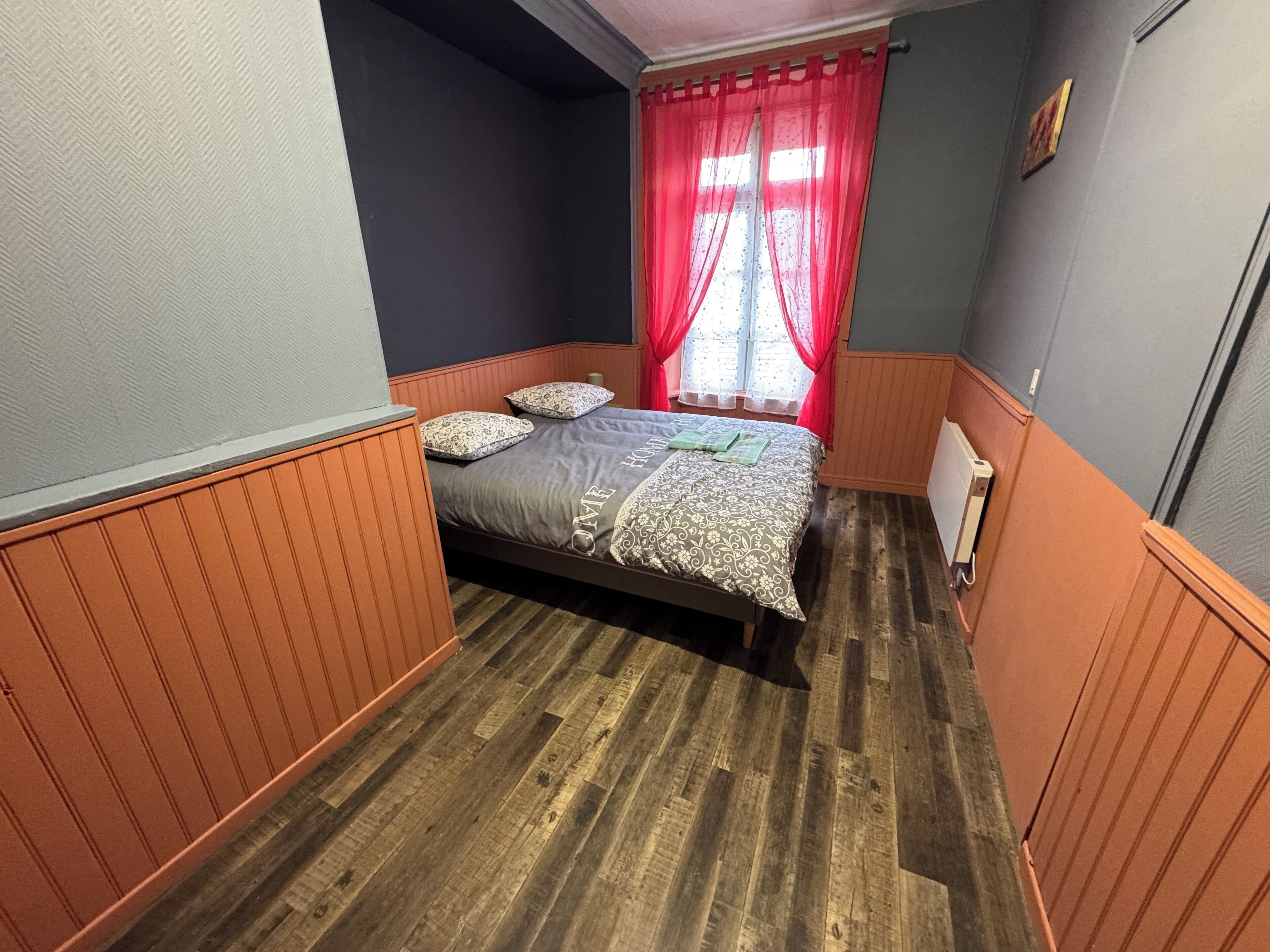 Een kamer met een bed, houten vloer, een radiator en een raam met rode gordijnen.