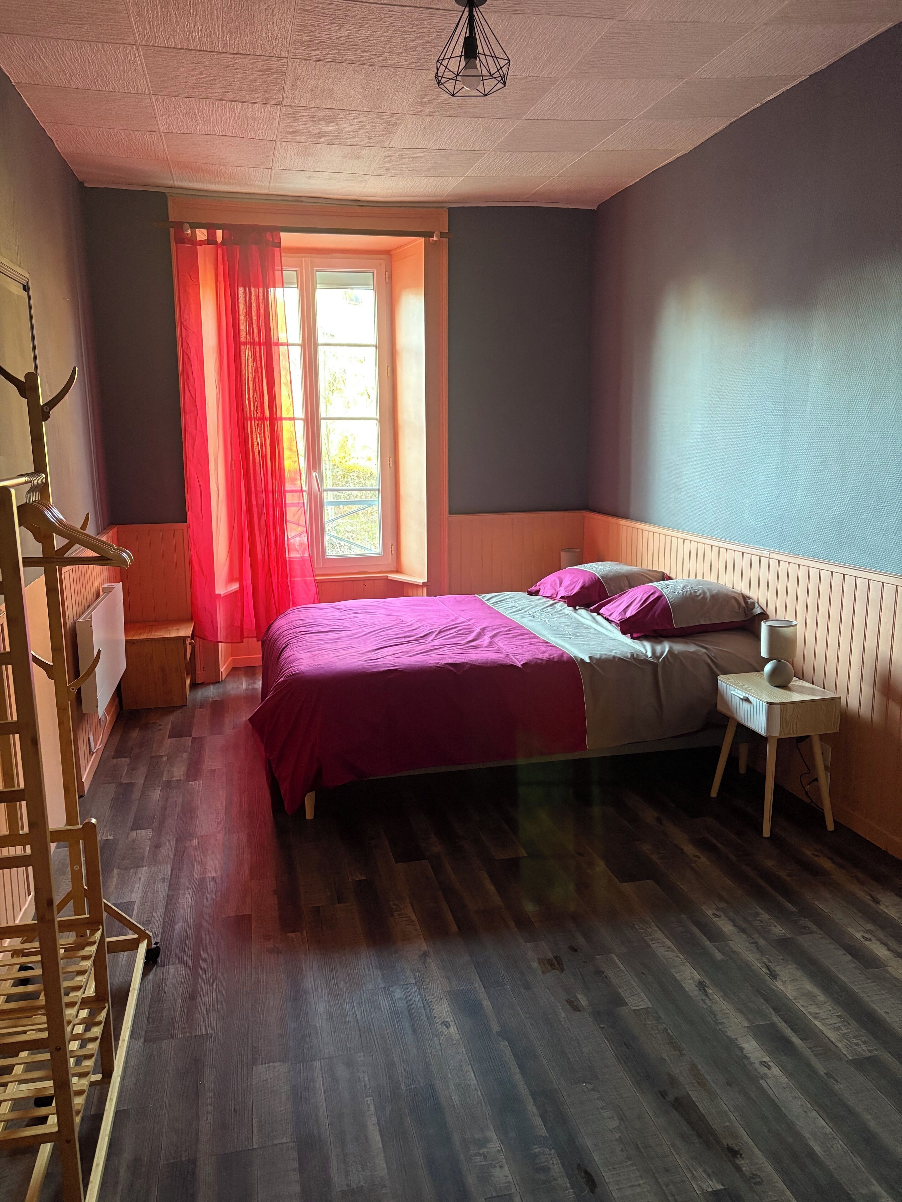 Et soveværelse med trægulv, en seng med lyserødt sengetøj, et natbord med lampe og et vindue med røde gardiner.