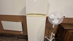 Ein weißer Kühlschrank mit Gefrierfach, ein weißer Ventilator und ein Holzkabinett in einem Raum mit Holzvertäfelung und dunklem Holzboden.