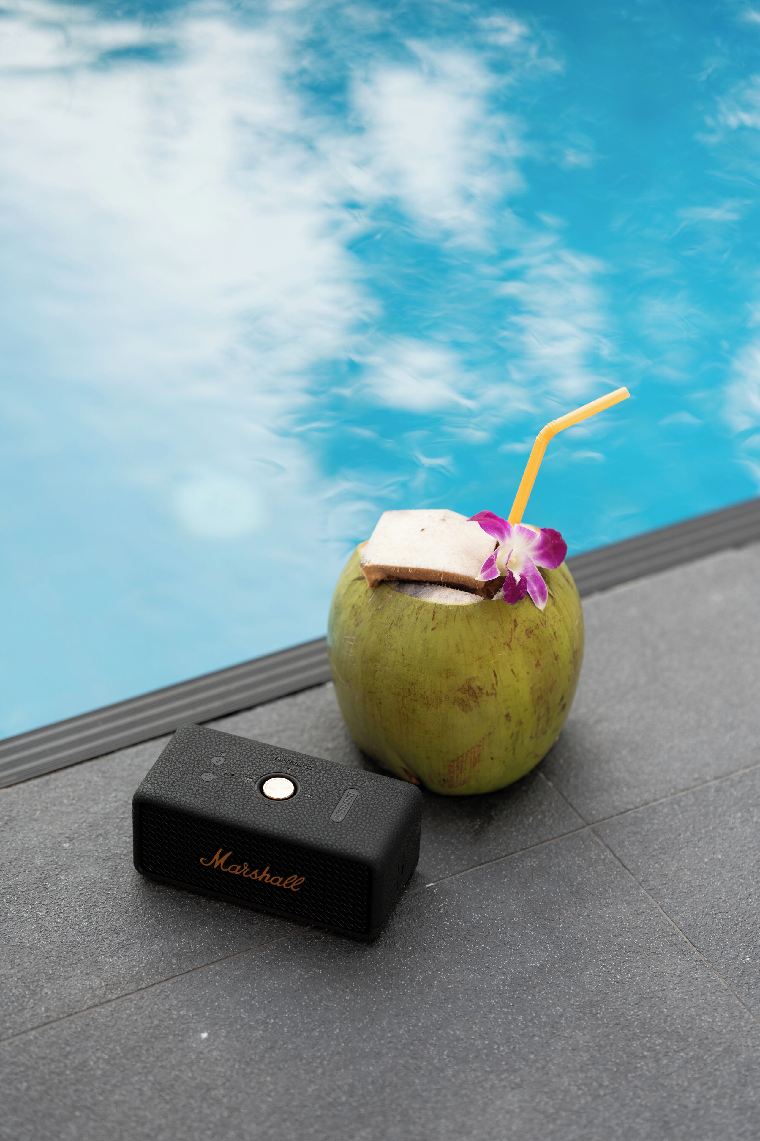 A green coconut with a straw and a piece of fruit on top, next to a black device labeled "Marshall" on a tiled surface by a pool.