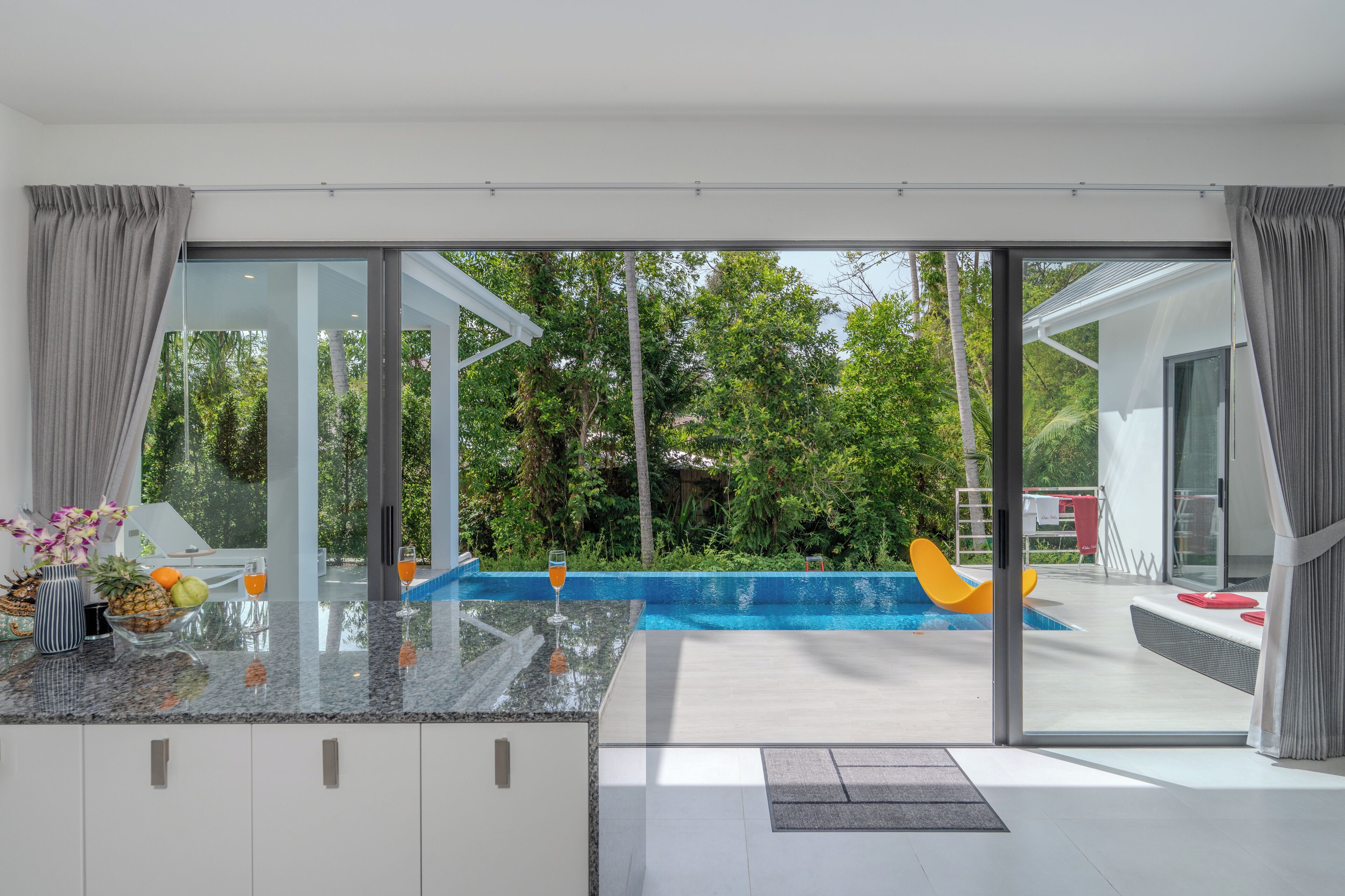 Eine moderne Küche mit einer großen Kochinsel, einer Glastür, die sich zum Poolbereich öffnet, und Blick auf üppiges Grün.