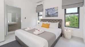 A modern hotel room with a large bed, a bathroom with a glass door, a painting on the wall, and a window with a view of greenery.