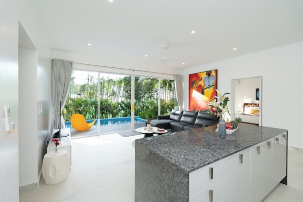 A modern kitchen with a granite countertop, a black leather sofa, and a large window overlooking a pool. - Osho Villas Koh Phangan (Ko Pha-ngan)