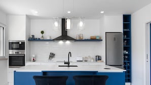 A modern kitchen with a central island, two bar stools, a built-in oven, a microwave, a stainless steel refrigerator, and a blue shelving unit. - Villa Serenity - a paradise between golf and the sea (Foz do Arelho)