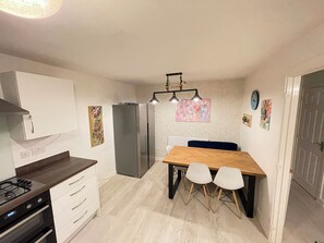 Eine kleine Küche mit Gaskochfeld, weißen Schränken, einem Kühlschrank und einem Esstisch mit zwei Stühlen.