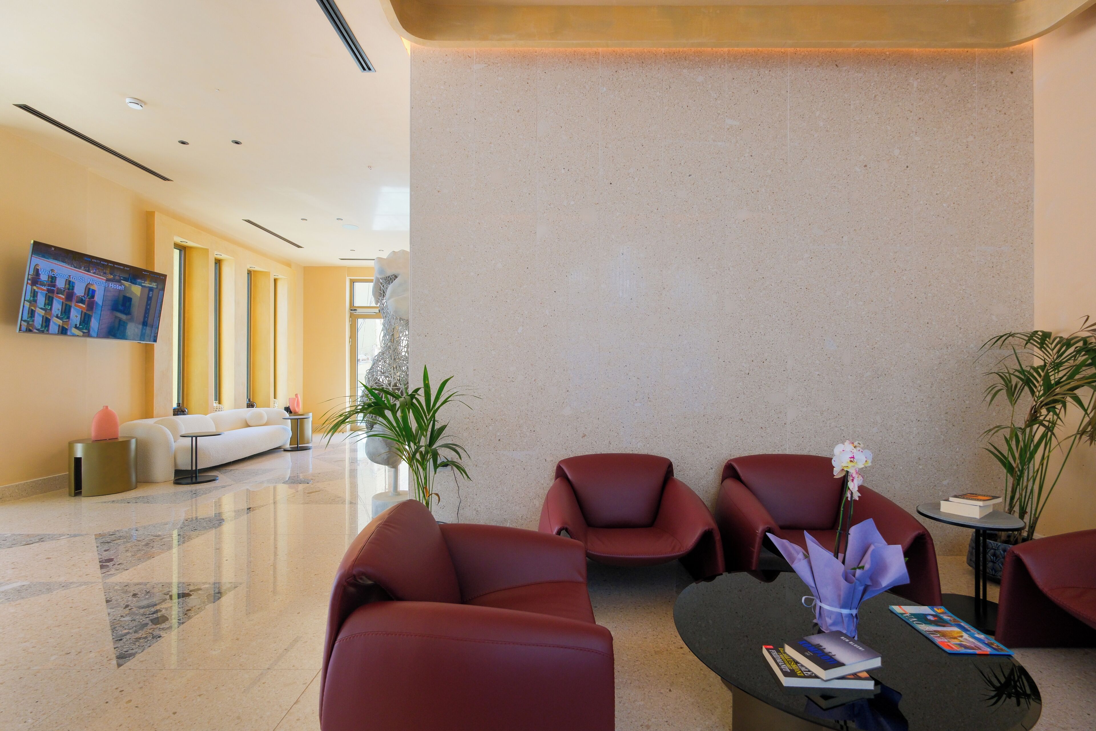 A modern lobby with red leather chairs, a black coffee table with books and a vase, and a large flat-screen TV on the wall.