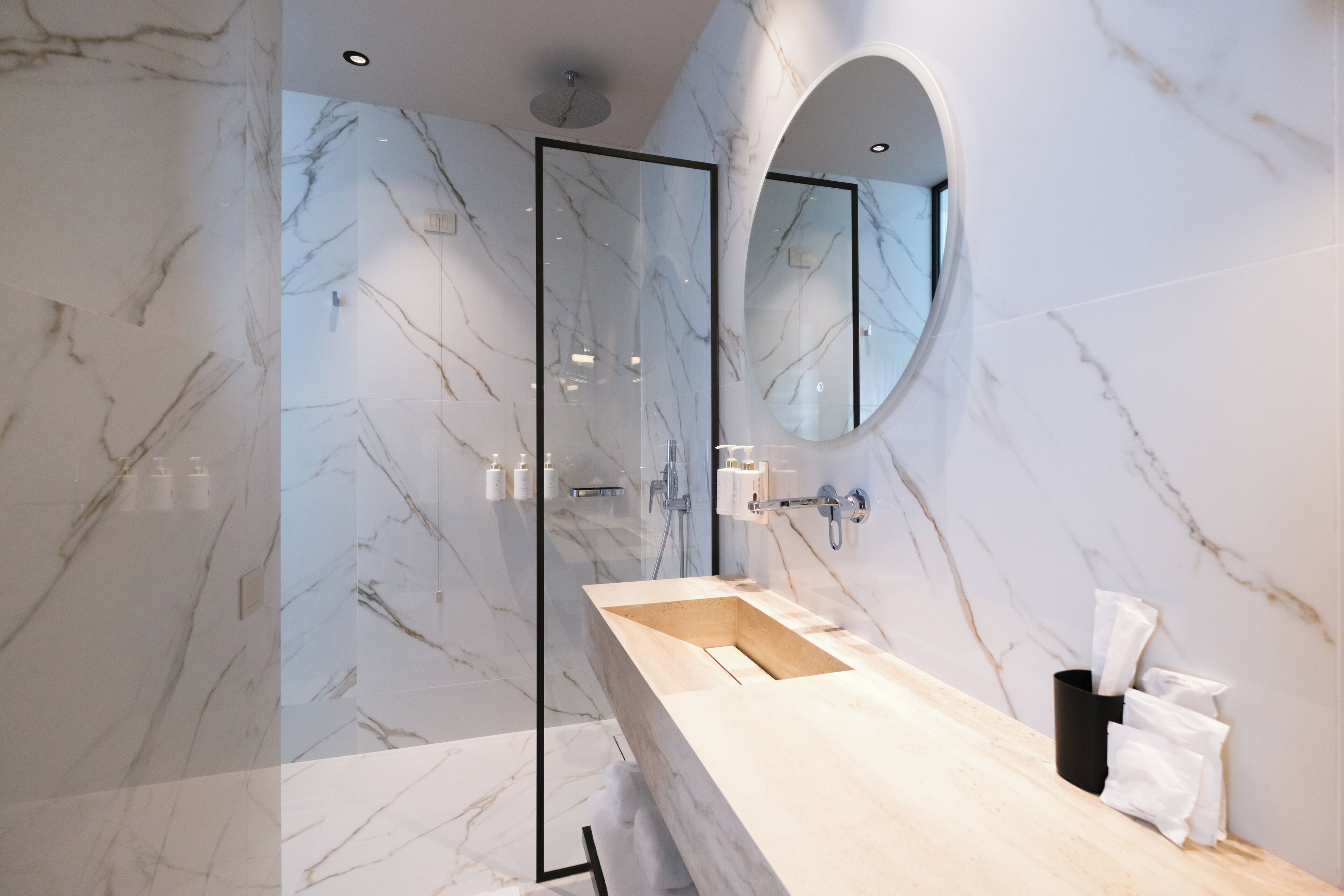 A modern bathroom with a marble wall, a wooden vanity, a glass shower enclosure, and a large mirror.