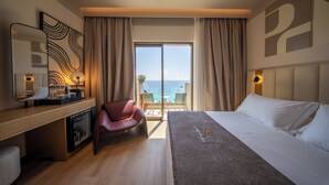 A hotel room with a bed, a desk with a mirror, a red chair, a minibar, and a view of the sea through large windows. - Saint Nicolas (Himarë)
