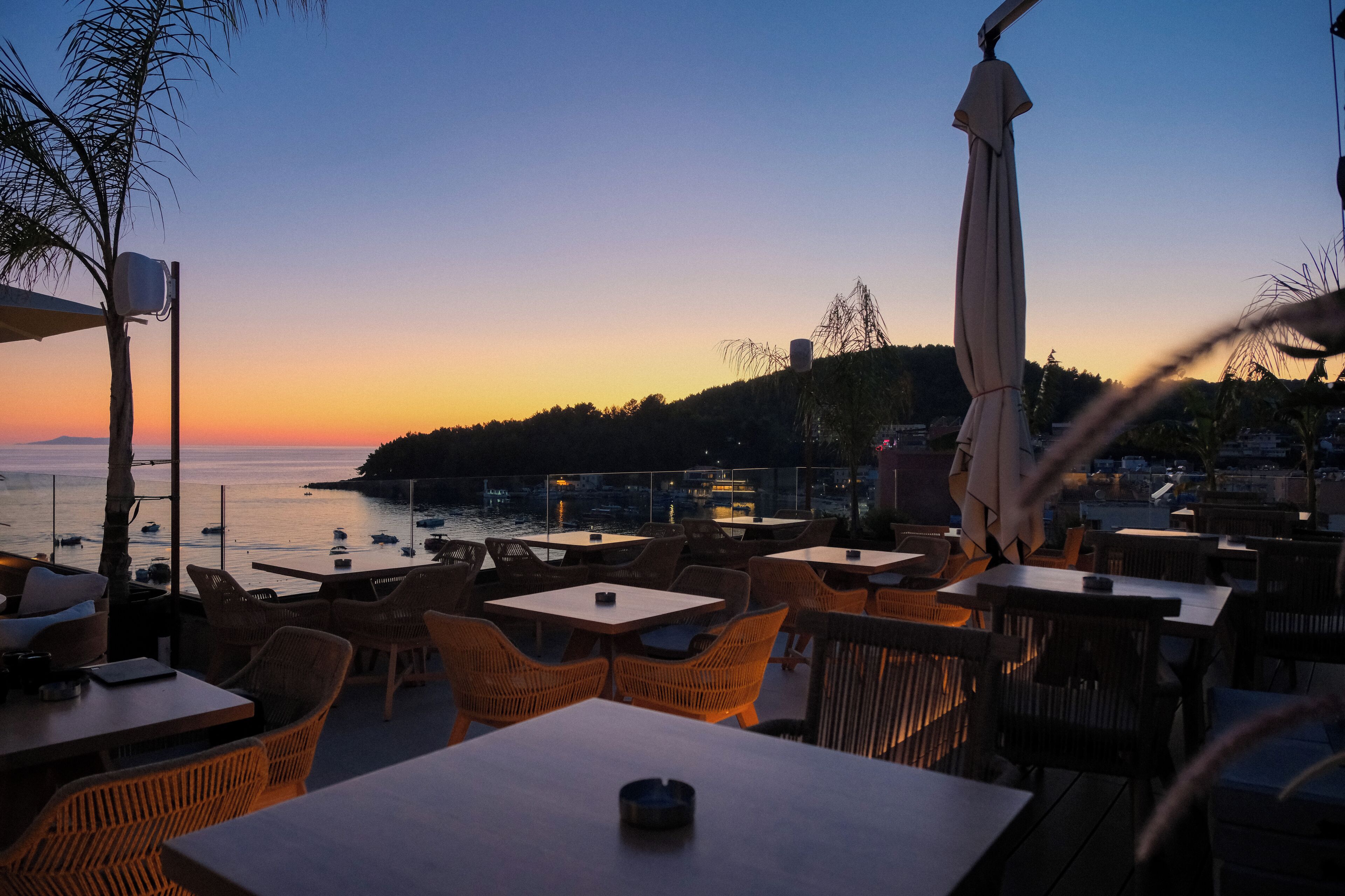 An outdoor seating area with tables and chairs, overlooking a body of water and a distant shoreline during sunset.