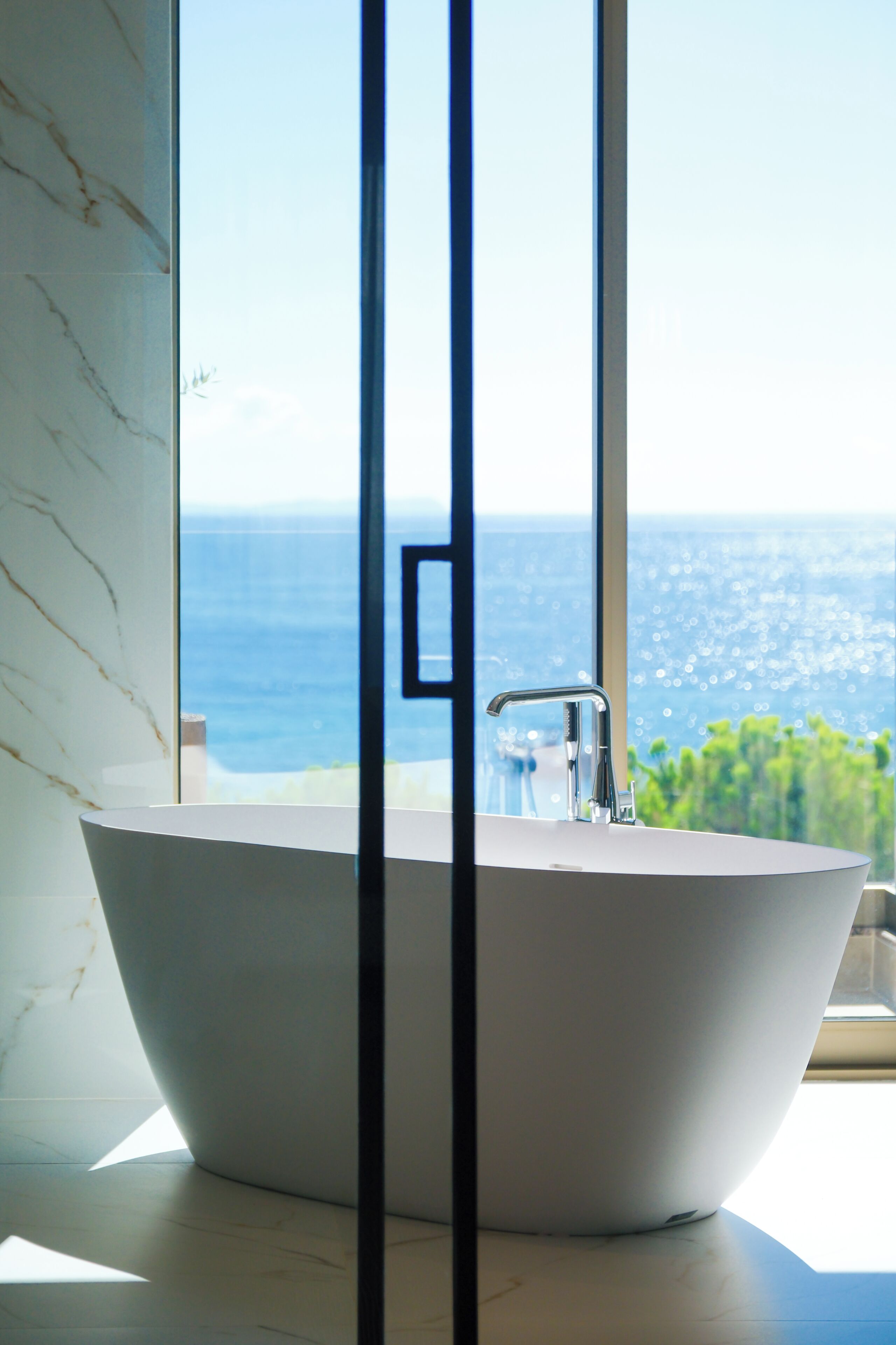 A modern bathroom with a freestanding bathtub, a large window, and a view of the sea.