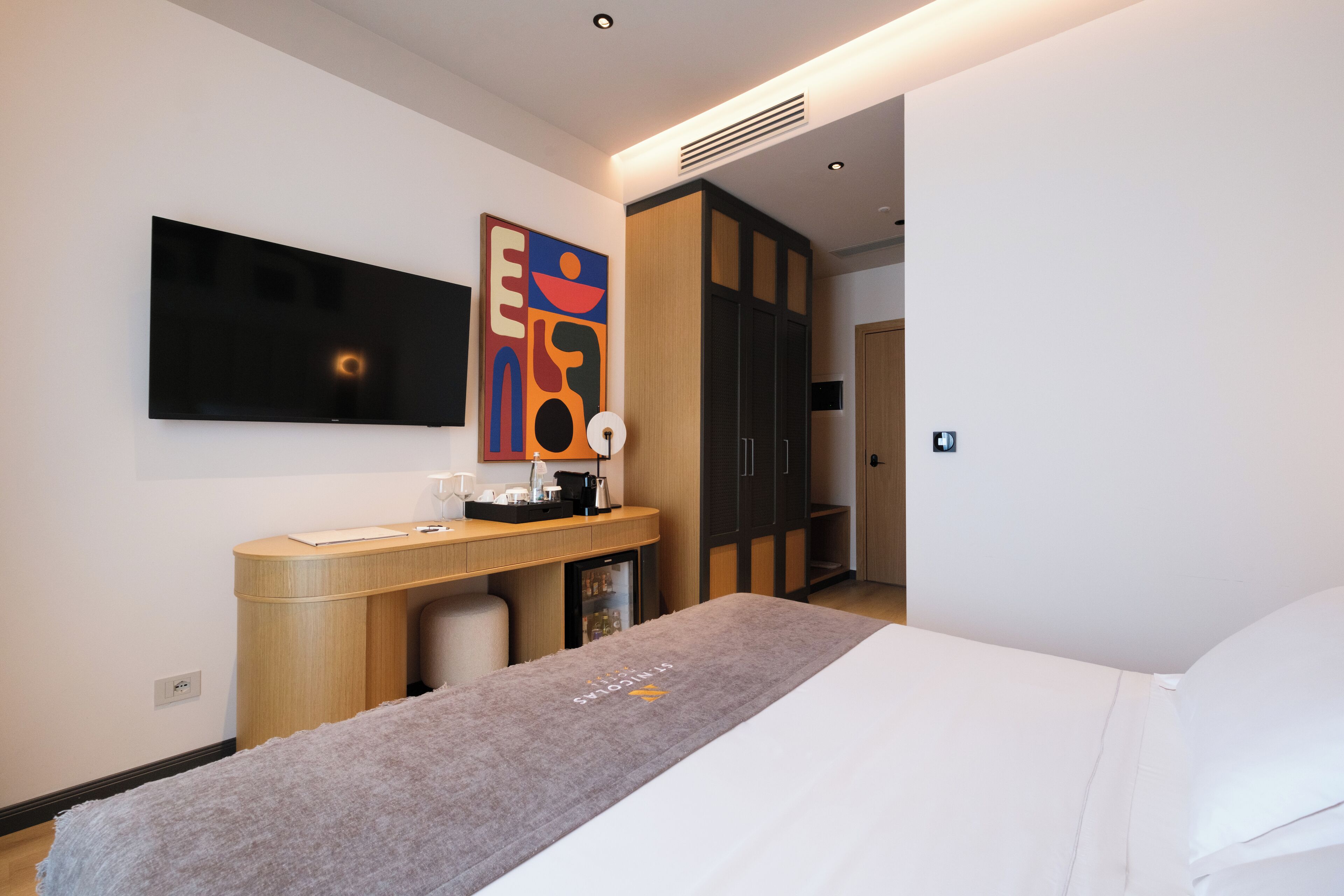A hotel room with a bed, a desk with a computer, a TV mounted on the wall, and a wardrobe.