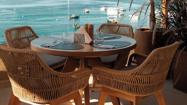 A round wooden table with wicker chairs, overlooking a coastal view with boats and a clear sky. - Saint Nicolas (Himarë)