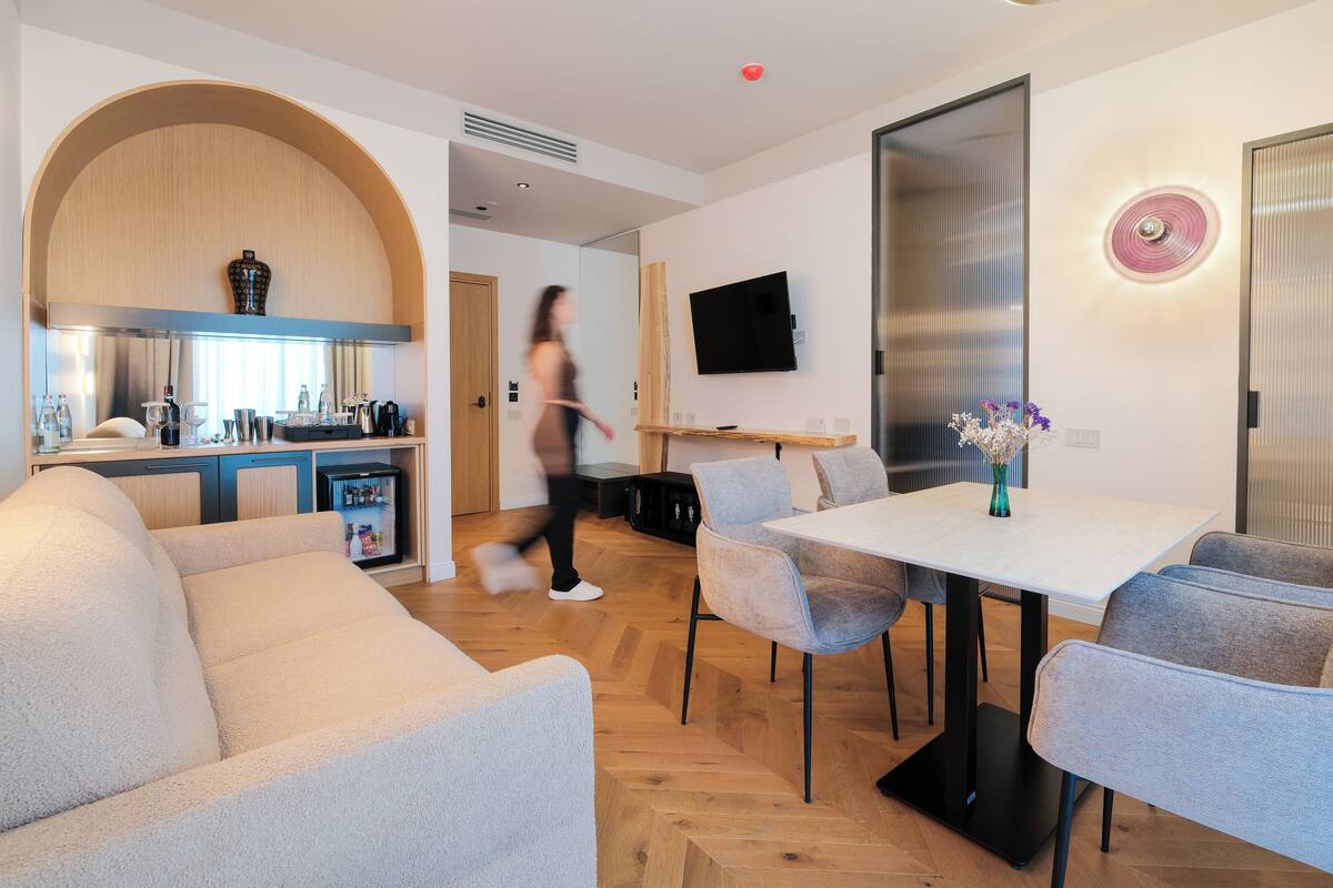 A modern living room with a sofa, a dining table with chairs, a kitchen area with cabinets and appliances, and a TV mounted on the wall.