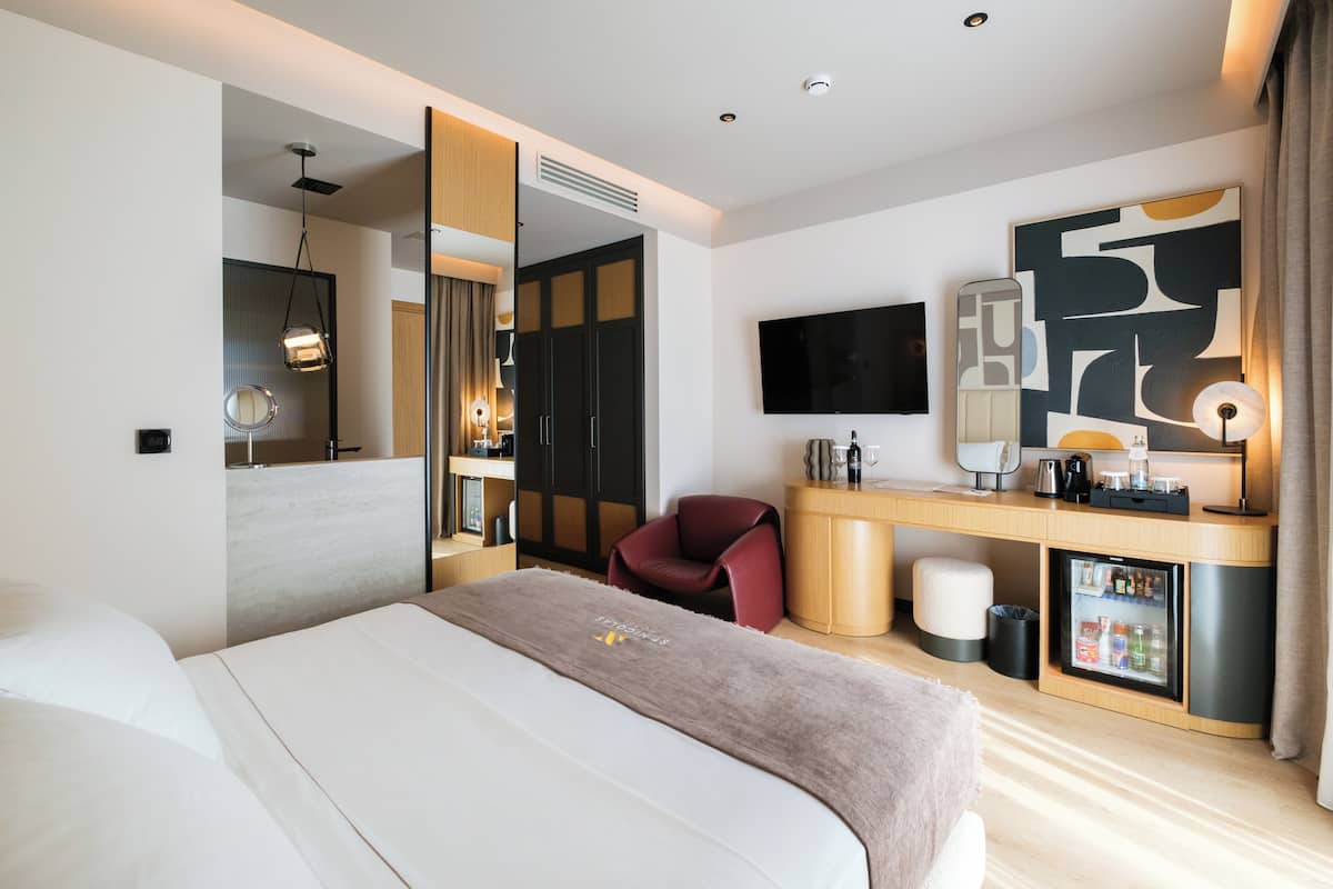 A modern hotel room with a large bed, a desk with a computer, a TV, and a minibar.