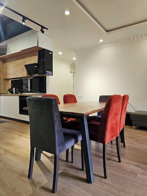 A modern dining area with a wooden table and four chairs, one grey and three red, in a well-lit room with a kitchen in the background.