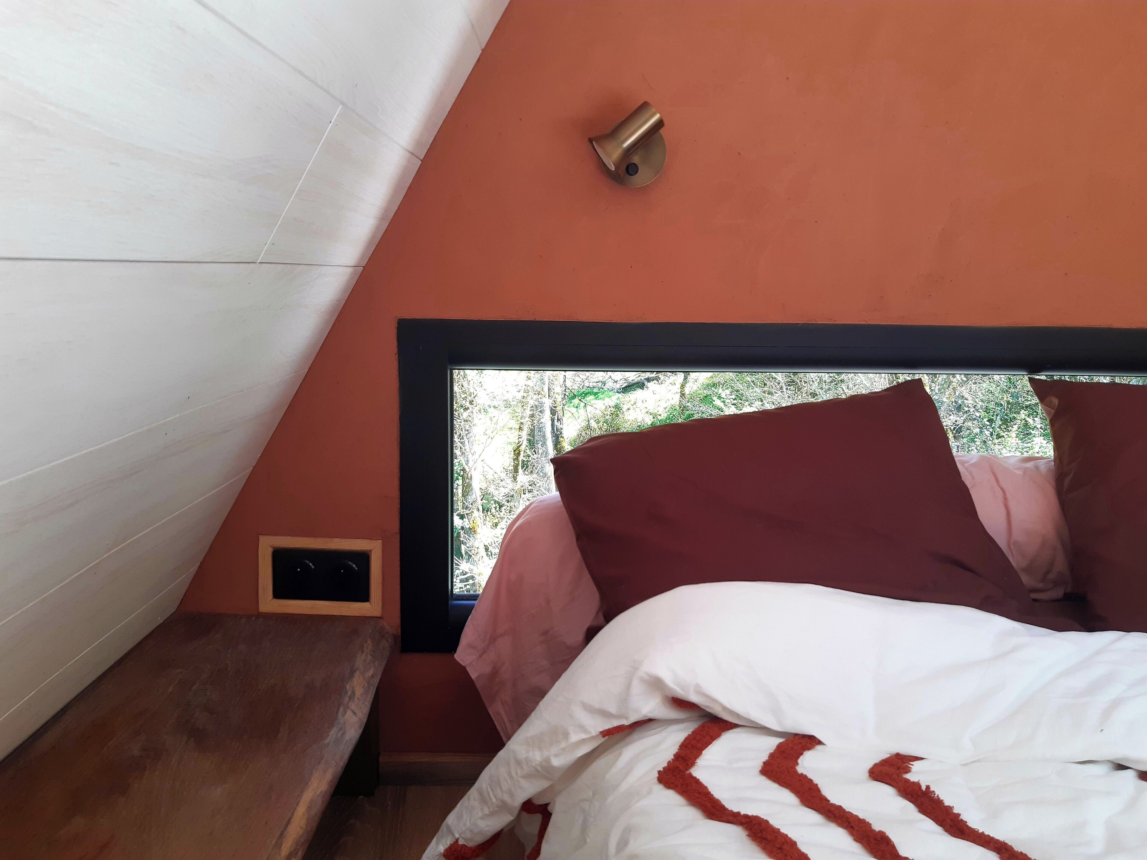 Une chambre avec un plafond en pente, une fenêtre donnant sur un paysage verdoyant, un lit avec une literie blanche et rouge, et une petite table de chevet.