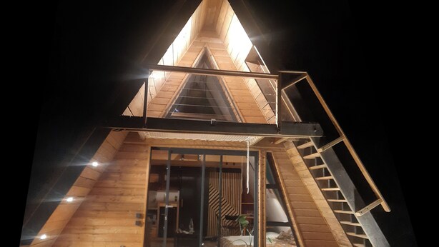 Een houten, driehoekige cabin met een glazen deur, een houten terras en een ladder naar het bovenste niveau.