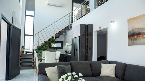A modern living room with a grey sofa, a coffee table, and a vase of white flowers. There is a staircase leading to an upper level with a view of the city. - Emma & Peris Villas-A Two Villa Retreat with Shared Courtyard,By IdealStay (Gouves)