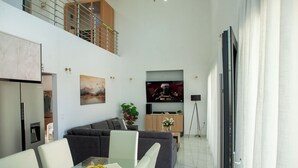 A modern living room with a glass dining table, grey sofas, a flat-screen TV, and a staircase leading to an upper level. - Emma & Peris Villas-A Two Villa Retreat with Shared Courtyard,By IdealStay (Gouves)