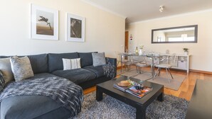 Un salon moderne avec un canapé gris foncé, une table basse et un coin repas comprenant une table et des chaises en verre. Des tableaux encadrés ornent le mur, et un grand miroir est placé au-dessus de la table à manger.