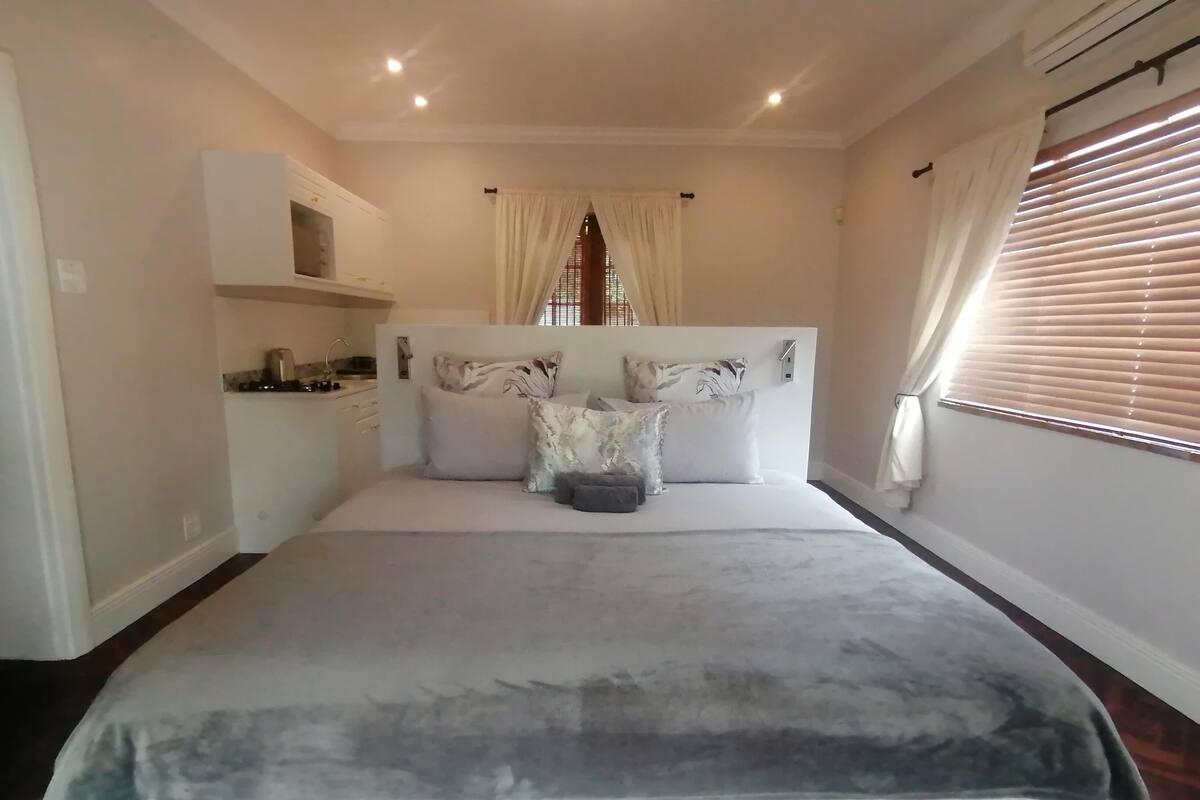A bedroom with a large bed, a built-in wardrobe, a small kitchenette, and a window with blinds.