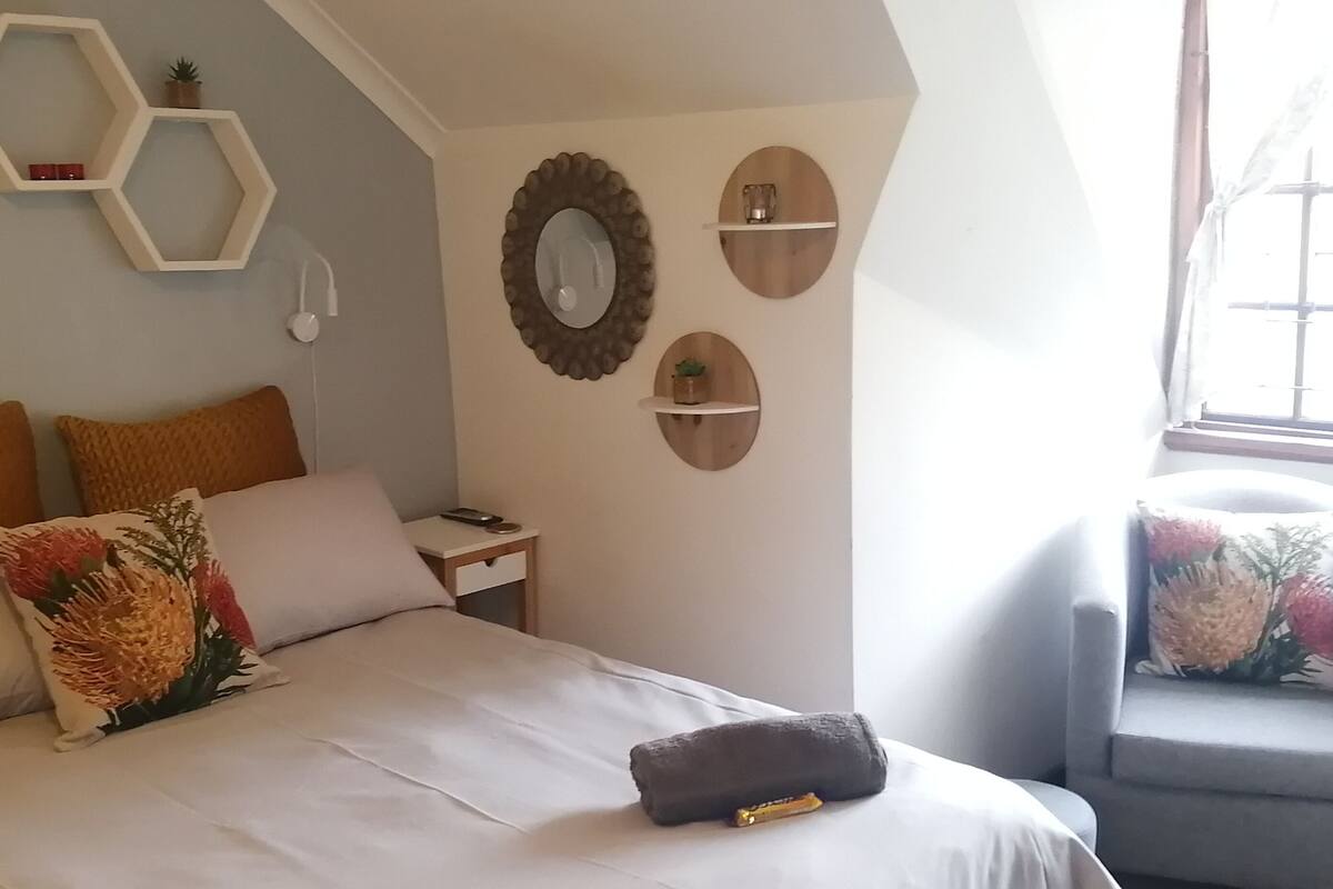 A bedroom with a bed, pillows, a bedside table, a chair, and wall decorations including hexagonal shelves and mirrors.