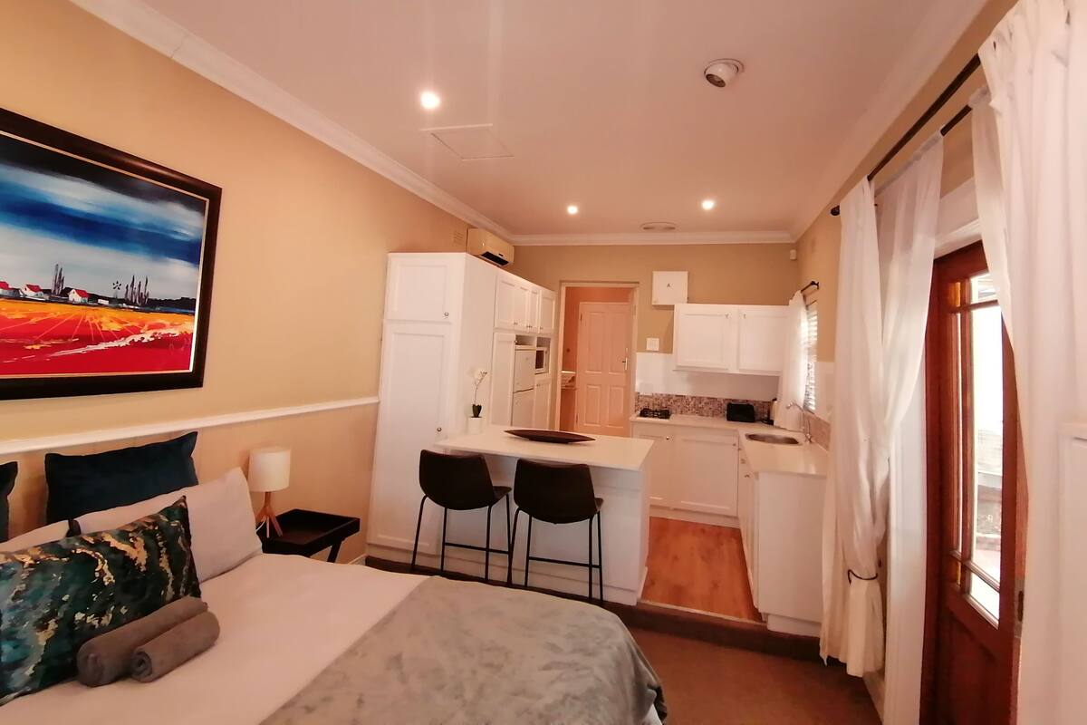 A compact hotel room with a bed, a desk, two chairs, a kitchenette, and a painting on the wall.