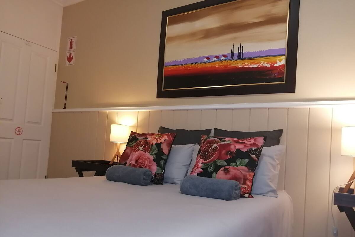 A neatly made bed with floral pillows, a framed painting above it, and a lamp on a bedside table.