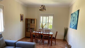A living room with a grey sofa, a dining table with chairs, a wooden cabinet, and a window with a view of greenery. - Chalet Villa Antonia (Ayamonte)