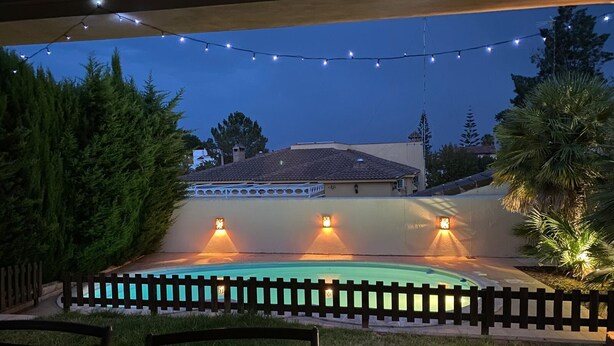 A swimming pool with a wooden fence, string lights, and a house in the background. - Chalet Villa Antonia (Ayamonte)