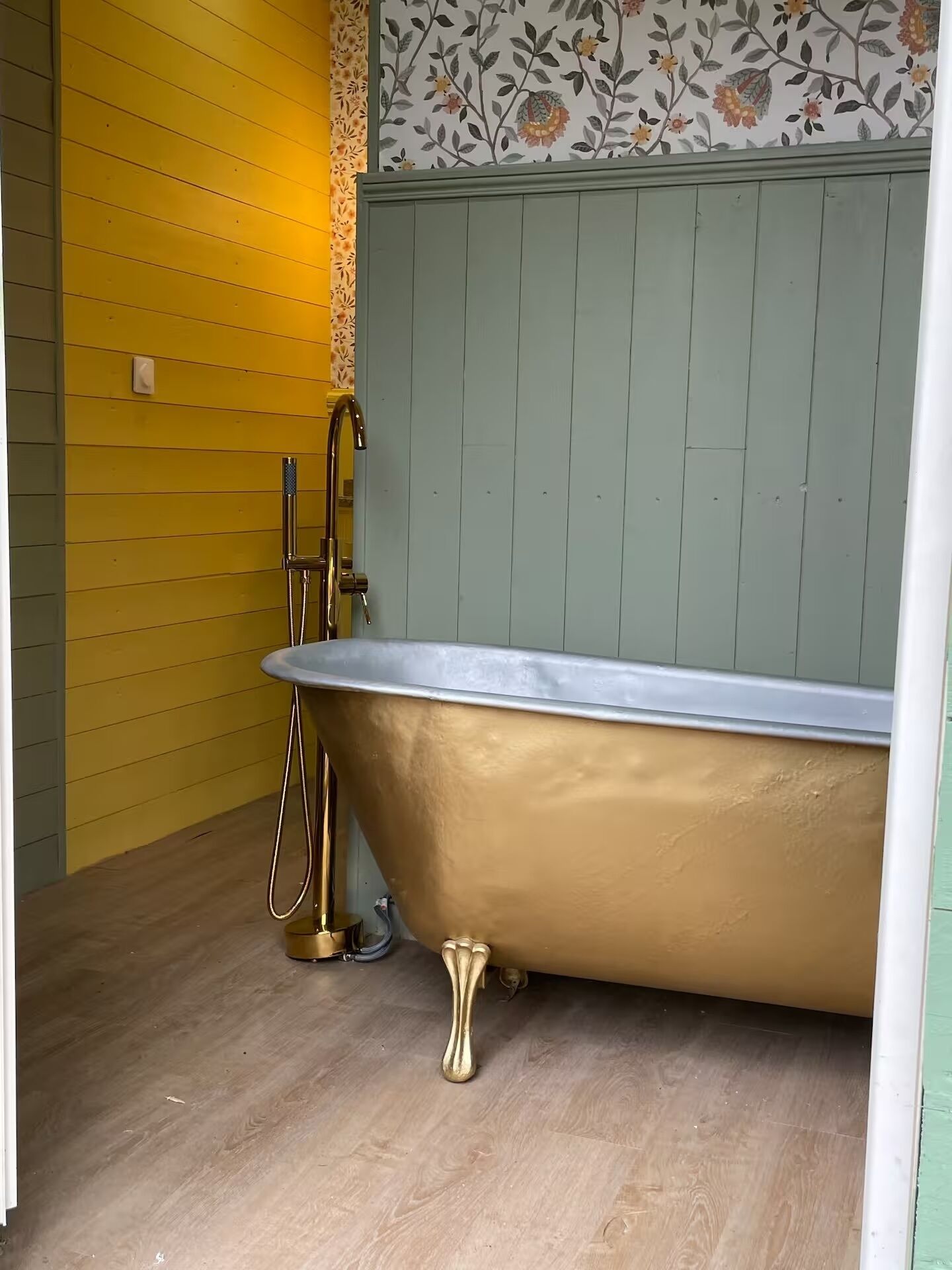 Een badkamer met een vrijstaand gouden bad, een gele wand en behang met bloemenmotief boven het bad.