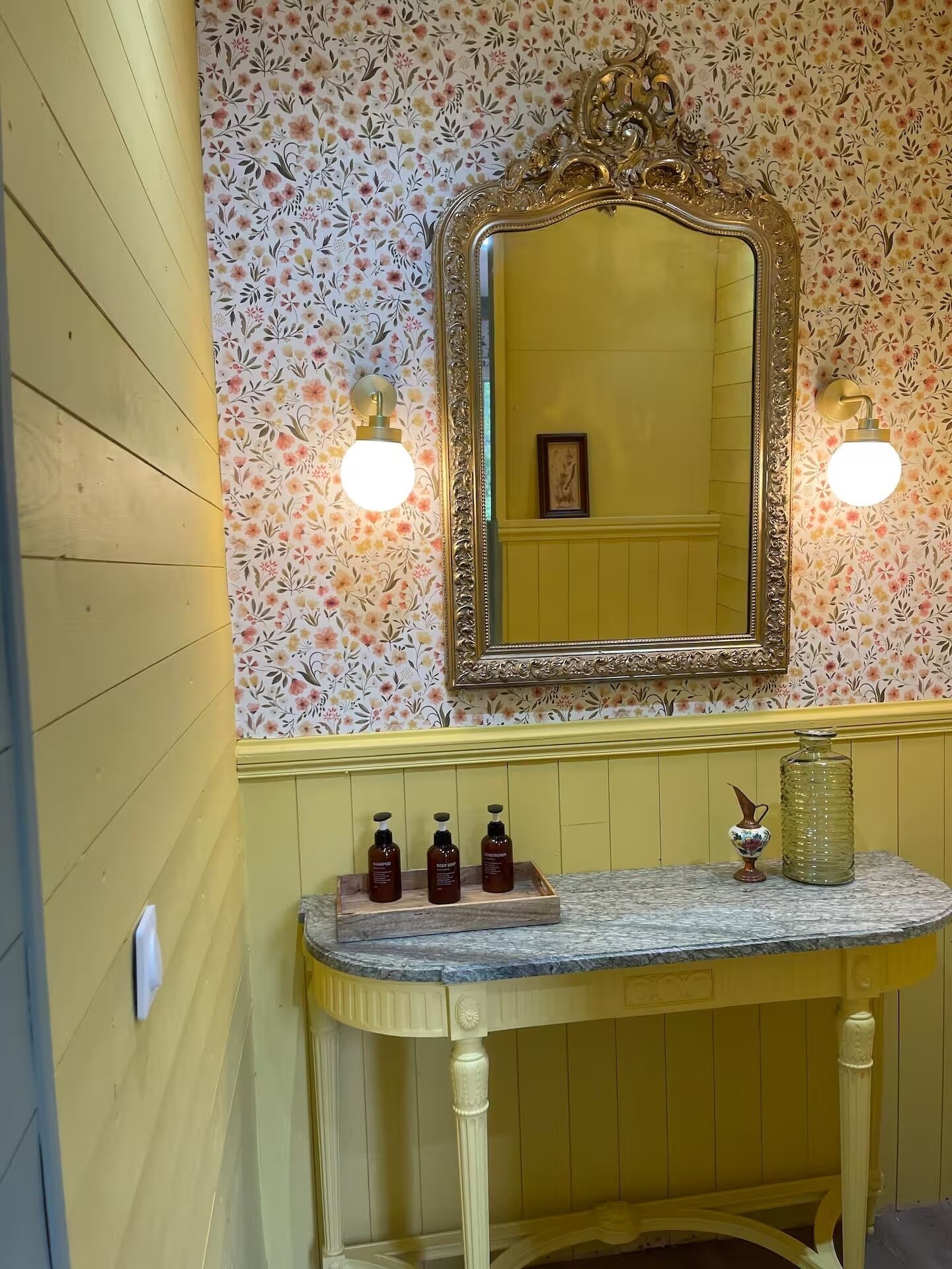 Un baño con papel tapiz floral, un espejo con marco ornamentado y un tocador con tres botellas y un jarrón.