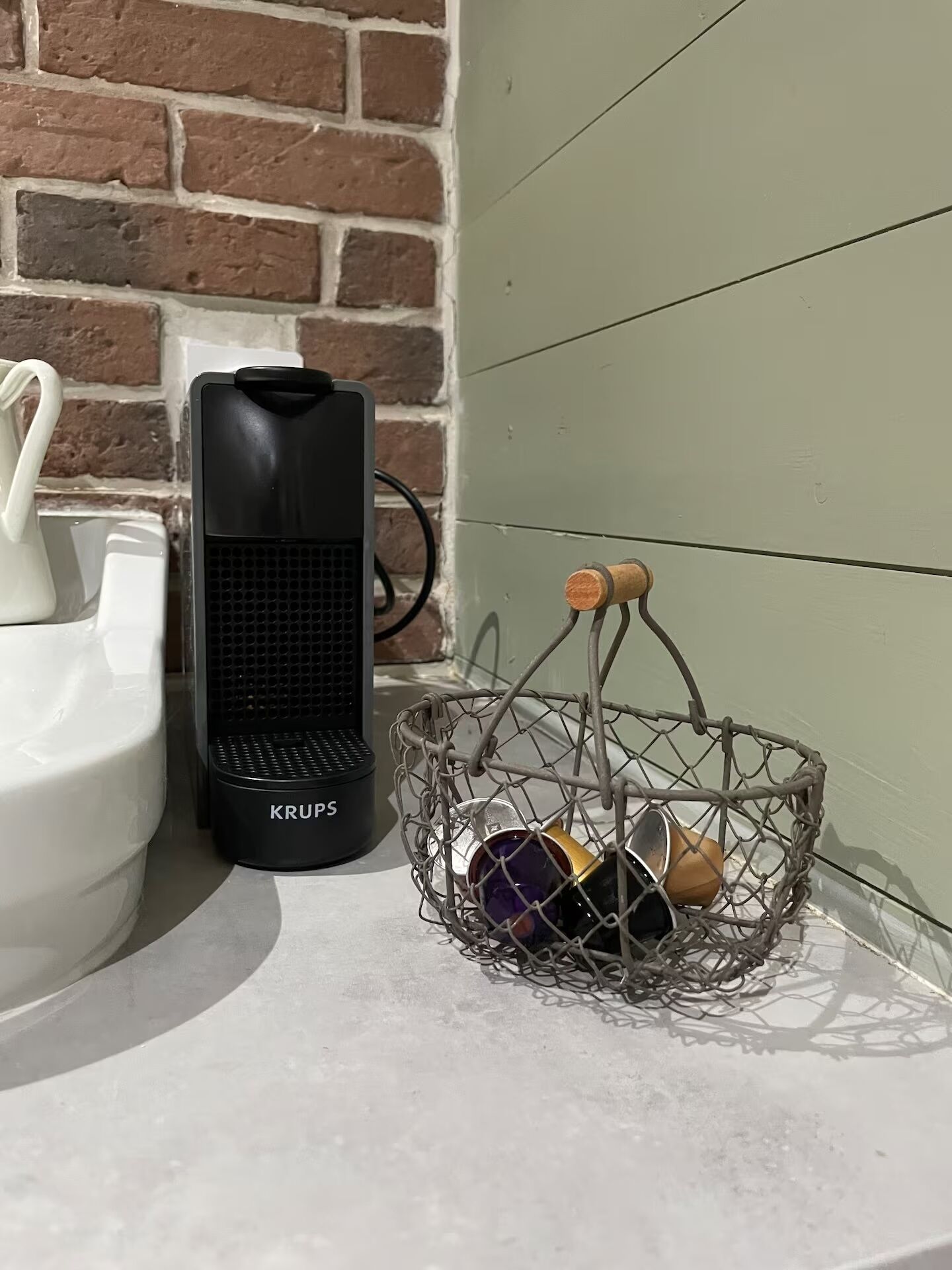 A bathroom with a white sink, a black Krups coffee maker, and a wire basket containing a purple mug and a wooden cup.