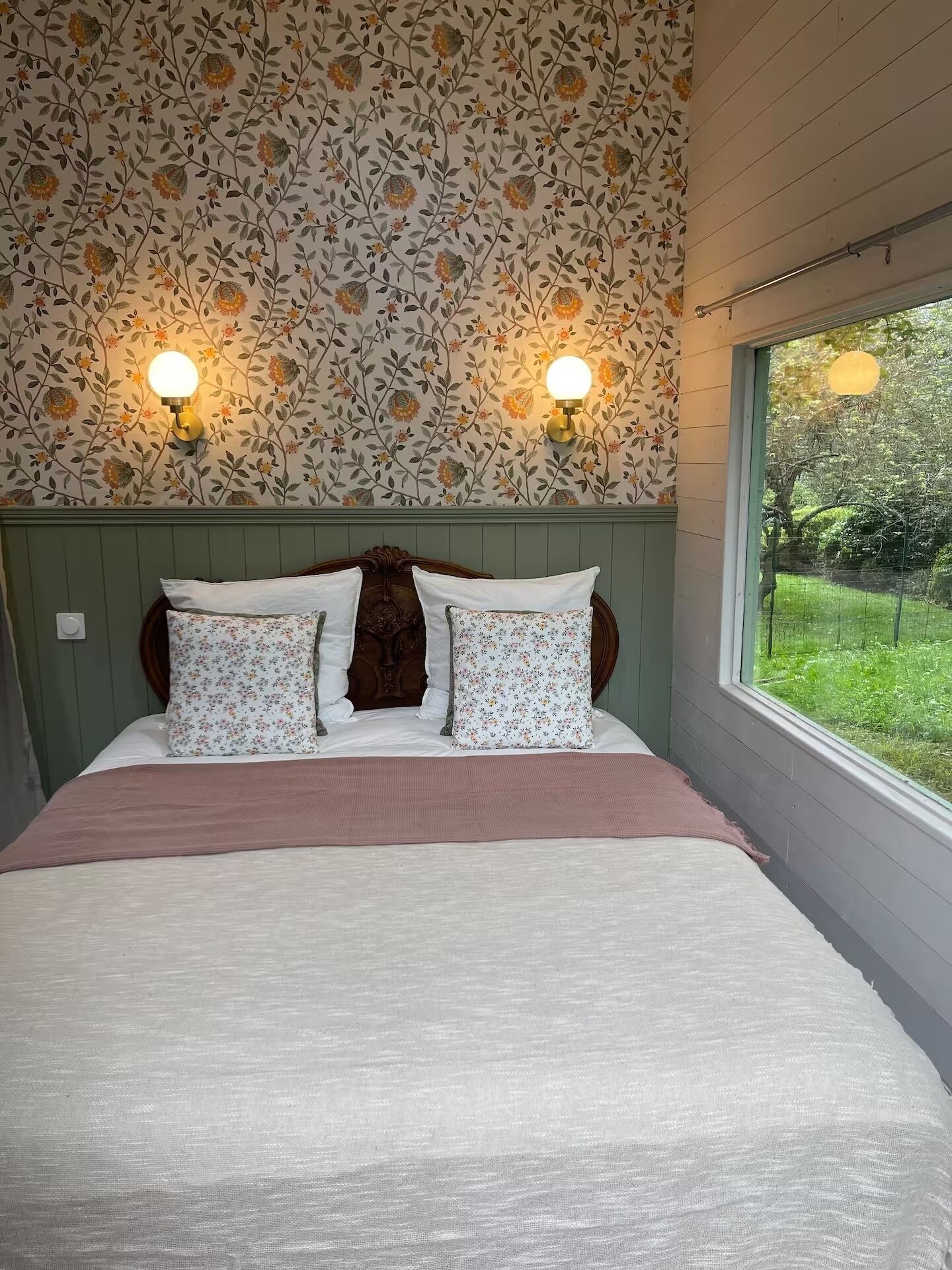 A bedroom with a floral wallpaper, two wall-mounted lights, a wooden headboard, and a view of greenery outside.