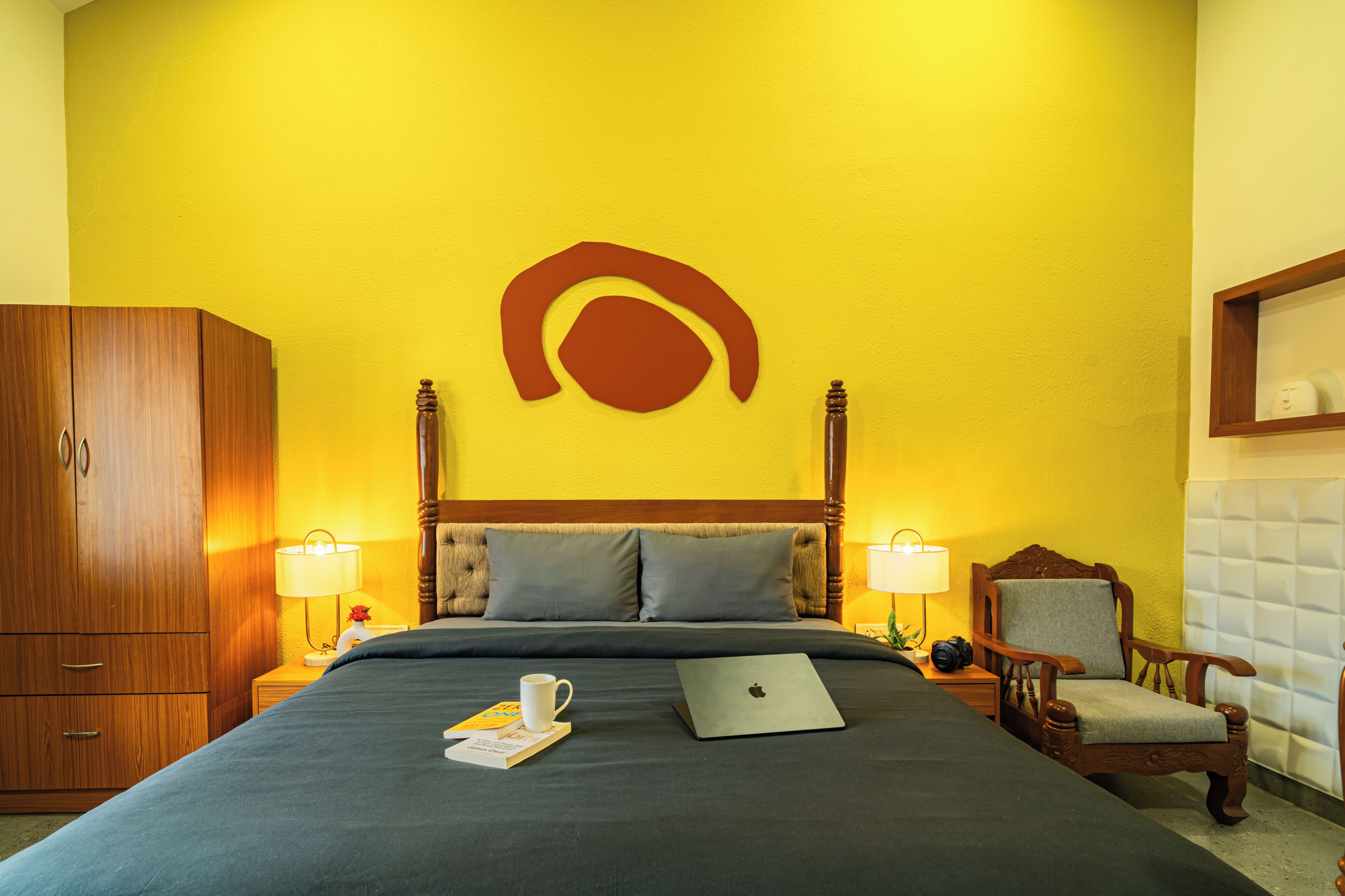 A hotel room with a bed, wooden wardrobe, two bedside lamps, a laptop, a chair, and a wall-mounted shelf.